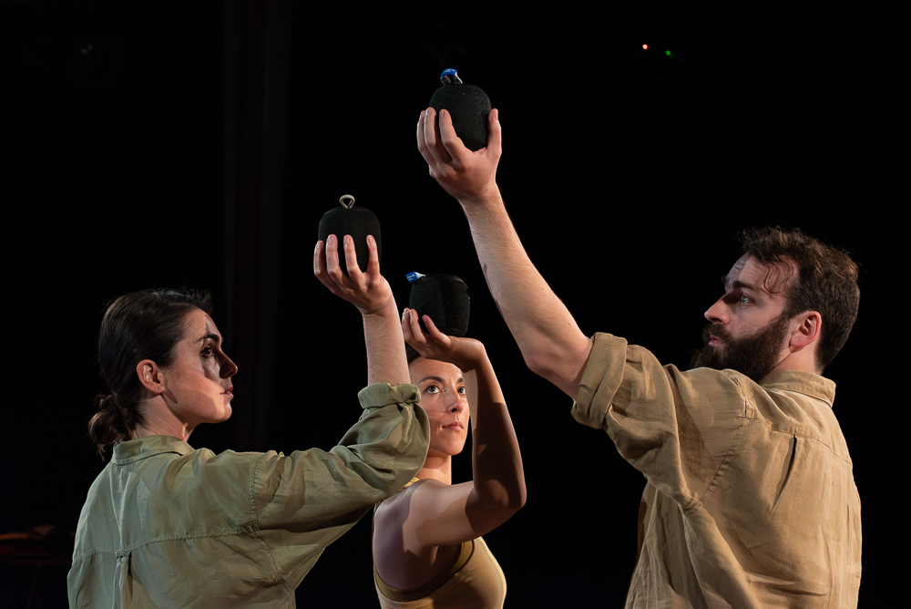 Three performers hold portable bluetooth speakers in the palms of their hands, their arms bent at the elbow, speakers raised just above their head.